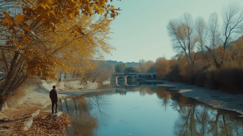 Man stranding along the shoreline of river, trees on either side, bridge crossing the river in the distance