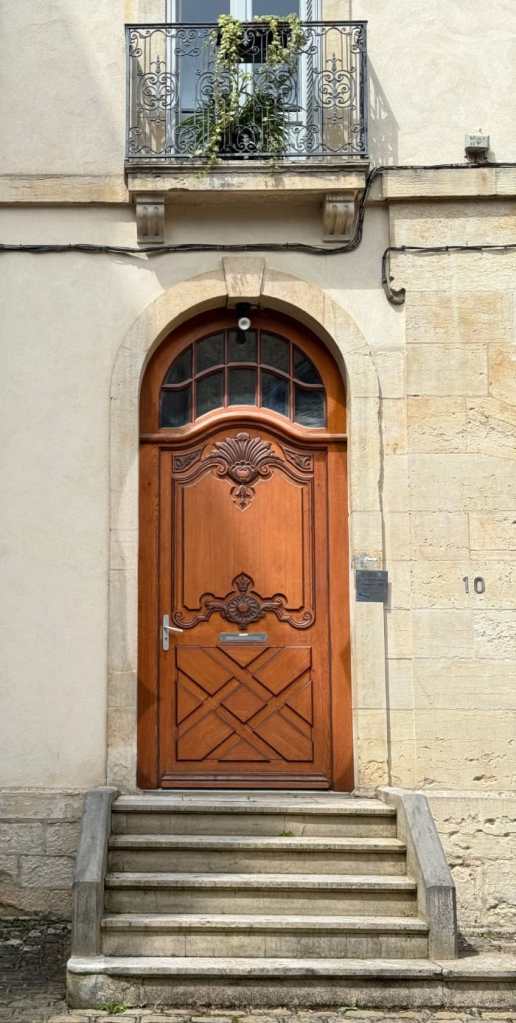 a wooden door with a distinct, classic design with a large, rounded top and intricate carvings. The carvings include floral and geometric patterns, adding a touch of elegance. The wood appears polished and warm in color. Above the door is a tall, arched window with smaller panes, allowing light to filter through. To the right of the door, there is a small metal plaque with an inscription, and next to that, a house number—10—is displayed. The door is framed by steps leading up to it, and the stone surrounding the door has a rough texture. A wrought iron balcony with greenery sits above the door, adding a touch of nature to the otherwise architectural scene. 