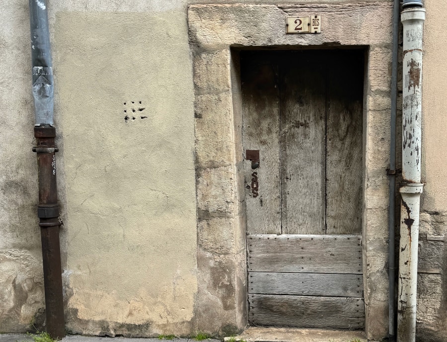 A weathered wooden door set deep within a rough stone frame, marked "2 bis" above, flanked by old metal drainpipes and a faded wall pierced with nine small ventilation holes in a grid pattern.