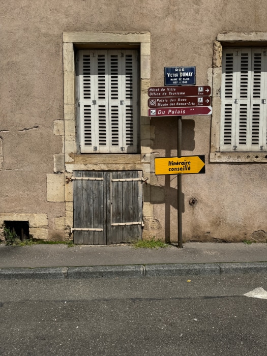This photograph captures a quaint, weathered street scene in Dijon, France. The composition centers around a rustic wall of a building with a muted beige facade, showing signs of age and wear. The texture of the wall is uneven, with cracks and patches that add character and suggest its historical nature.