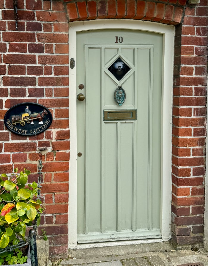 a sage green door with a diamond-shaped spyglass hole and a grass knocker with the letter slot mid-door. The plaque by the door names the brick cottage as "Brewery Cottage"