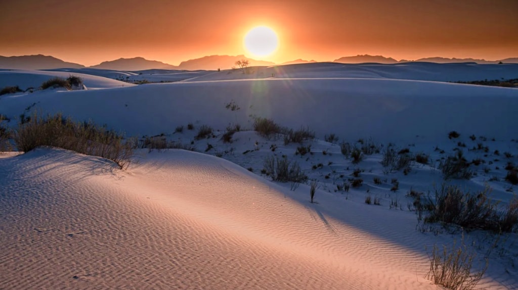 The sun is setting over a snowy landscape. A stunning sunset over the vast sand dunes, casting a gentle glow on the rippling sand and sparse vegetation.