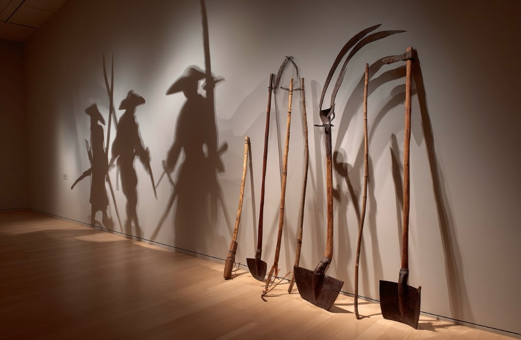 farm tools leaning against a wall in a museum with shadows of imaged farm workers reflected on the wall