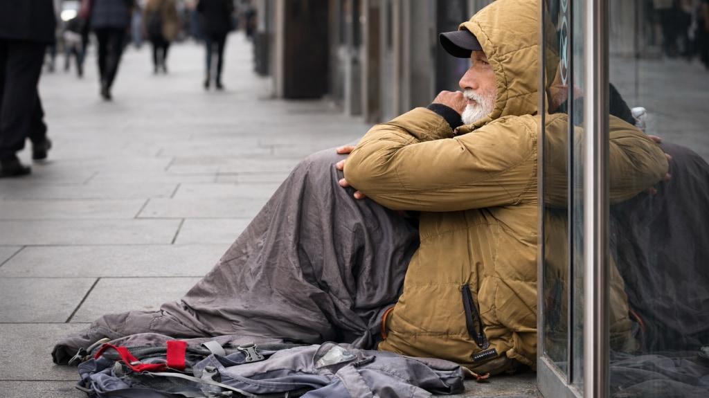 An elderly homeless man sits on a London pavement against a shop window, his belongings gathered at his feet as blurred pedestrians pass by.