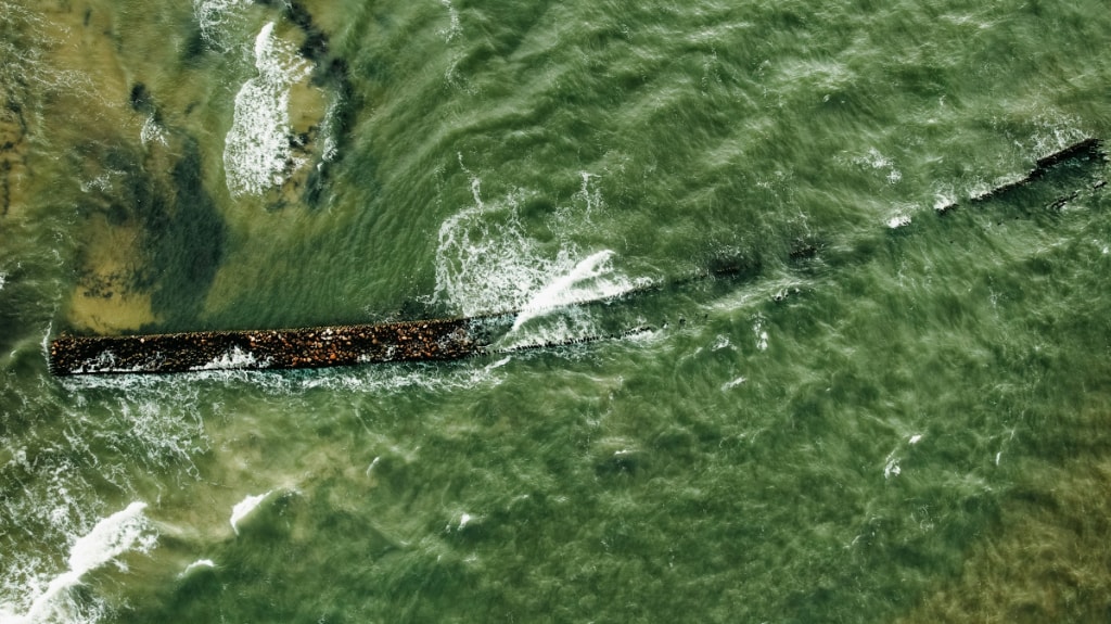 an aerial view of waves crashing over a submerged shipwreck at sea. The water shows us varying hues of colour, but primarily green.